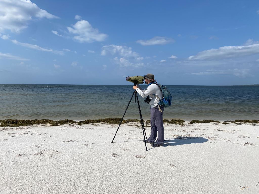 A man looks through a spotting scope while standing on a beach.